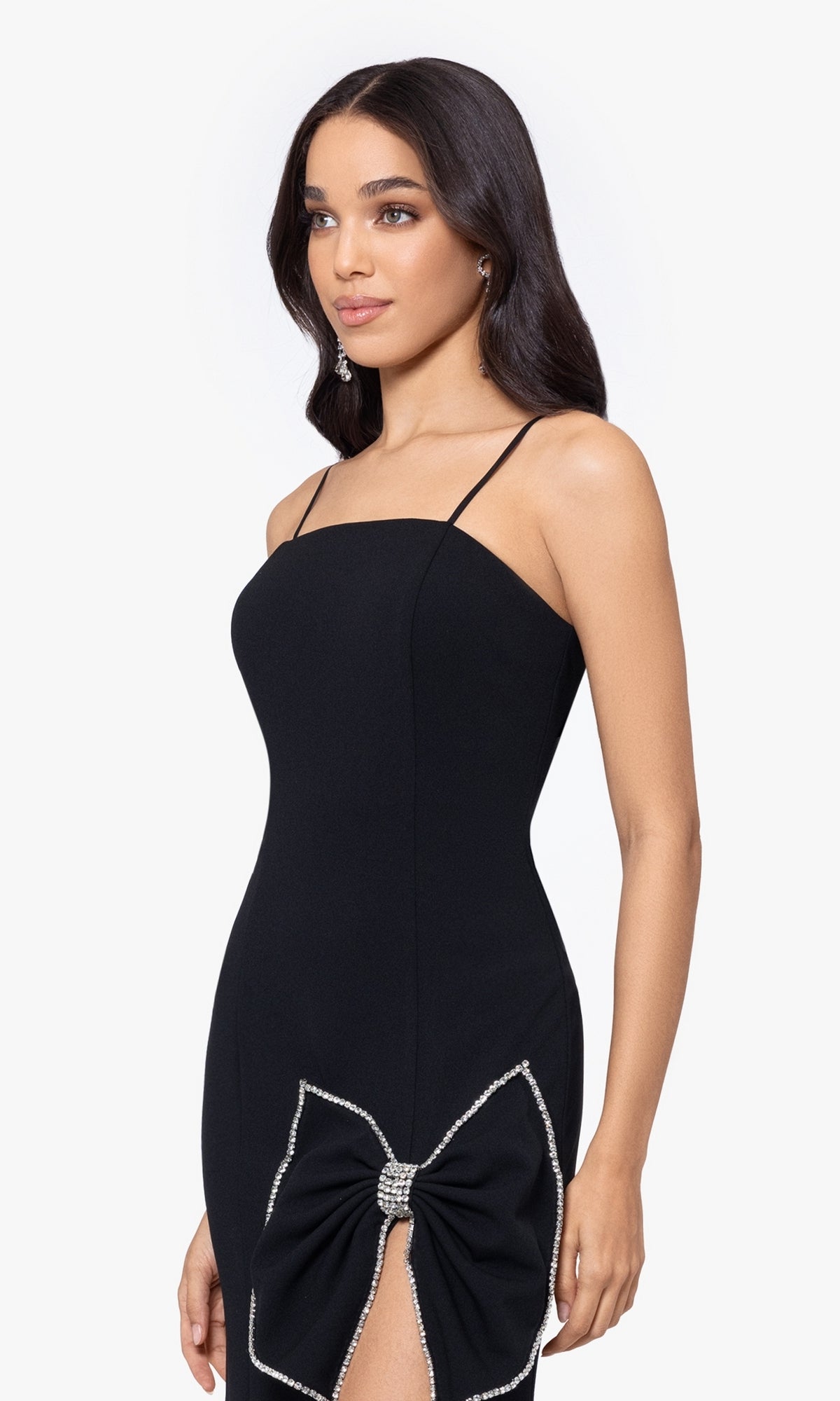 Woman wearing the Long Prom Dress 5063BN by Blondie Nites, a fitted black formal dress with thin straps and rhinestone bow detail at the hip, standing against a plain white background.
